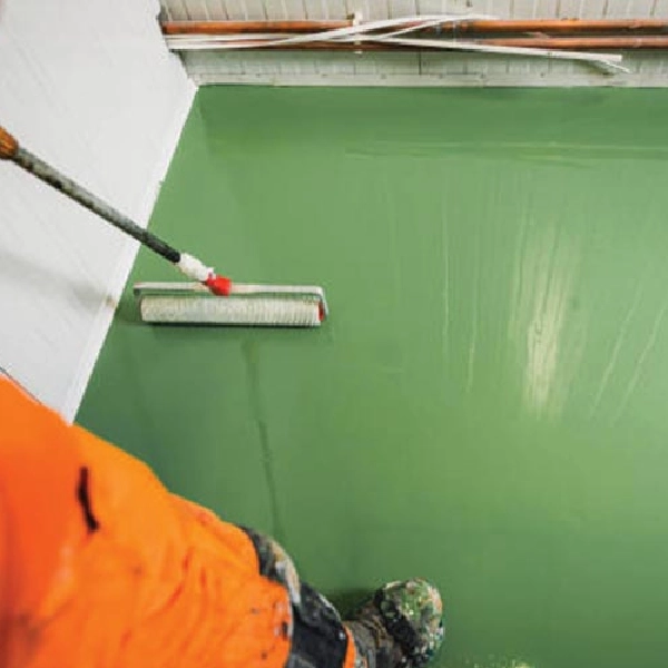 A person in orange work pants is seen from above, applying a vibrant green epoxy or floor paint to a concrete surface using a long-handled roller.